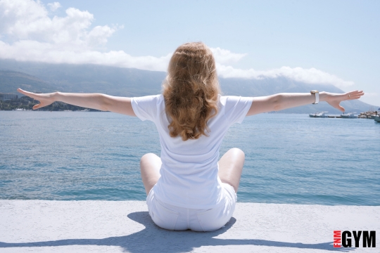 Woman sitting looking over the water with arms out working on her wellness